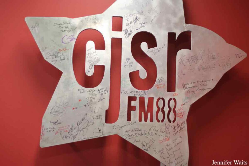 Photo of star-shaped silver color metal sign with the letters "c j s r" carved out of the center. Signatures are dispersed throughout the sign, which is hung on a red wall. Photo: J. Waits