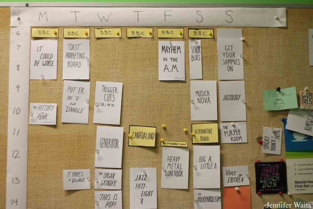 Photo of radio station schedule at CJSR-FM. Schedule is affixed to a bulletin board with push pins and each program name is handwritten on a rectangular piece of paper. Photo: J. Waits