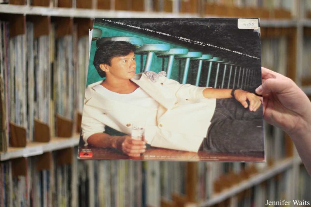 Photo of album cover held in front of record library at radio station CJSR-FM. Album cover art features a photo of a man laying down and holding a class of water. Photo: J. Waits