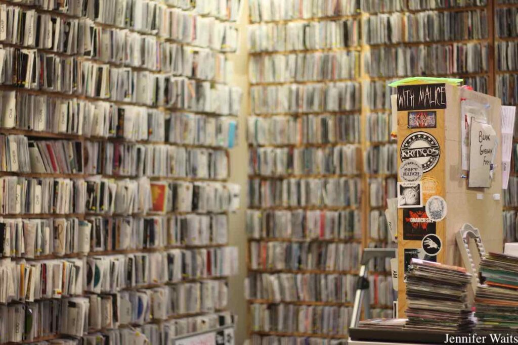 Photo of CD library at campus and community radio station CJSR-FM. Walls are covered with shelves packed with CDs. In the foreground, there are stacks of CDs and a shelf with stickers affixed to it. Photo: J. Waits