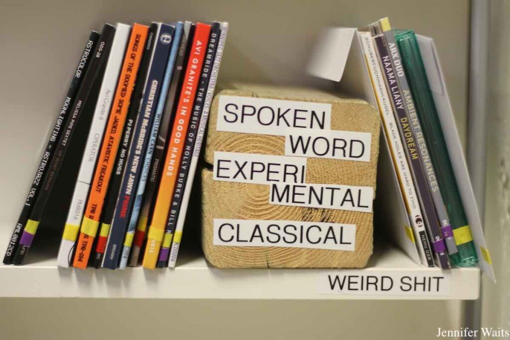 Photo of CDs on a shelf, with wooden block in the center of the frame. The block is labeled "spoken word experi mental classical" and the shelf is labeled "weird shit." Photo: J. Waits