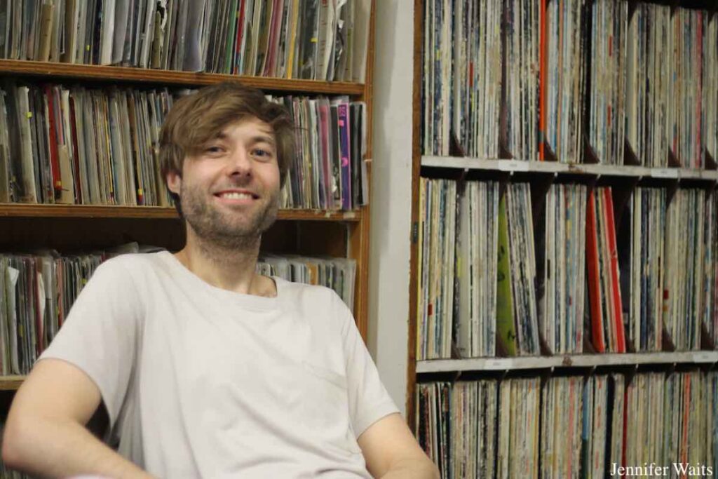 Photo of man wearing white T-shirt sitting in front of a wall of shelves filled with vinyl records at campus and community radio station CJSR-FM. Photo: J. Waits