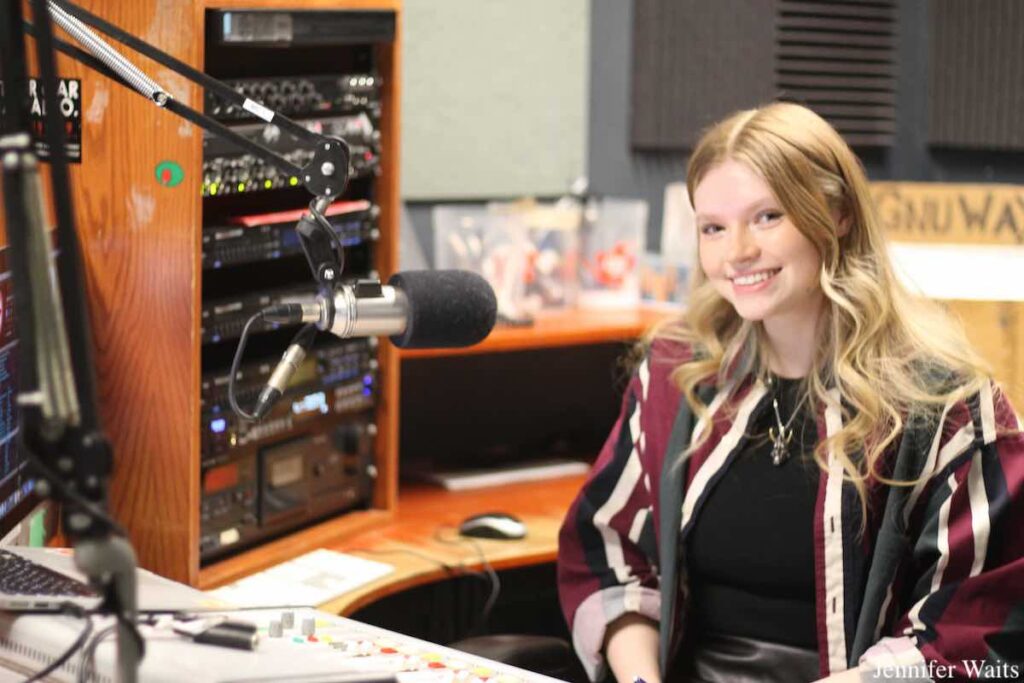 Photo of DJ sitting in on-air studio at campus and community radio station CJSR-FM. A rack of audio equipment can be seen, with a microphone in the foreground. Photo: J. Waits